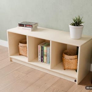 May include: A light-colored wooden storage unit with three open compartments. The top surface holds a white potted plant and two stacked books. Inside the cubbies are two woven baskets and a row of books titled Colorblind, Lost Souls.