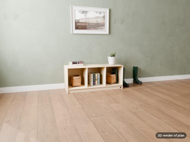 May include: A light-colored wooden storage unit with three open cubbies, containing two woven baskets and a stack of books. The unit sits on a light wood floor against a sage green wall, with a framed picture above and dark green boots nearby.
