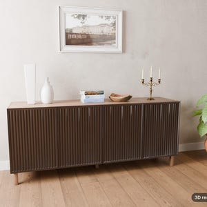 May include: A long, dark brown sideboard with a vertical ribbed texture on the four cabinet doors, supported by light-coloured wooden legs. The top surface holds two white vases, a stack of books, a wooden bowl, and a gold-toned candelabra.