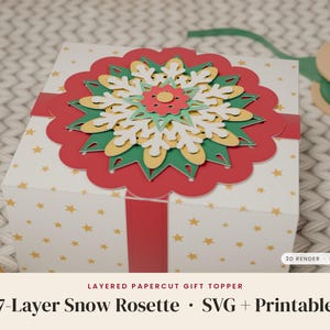 May include: A white gift box with gold stars features a layered papercut snow rosette topper in red, green, yellow, and white. A red ribbon wraps around the box. Text on the image reads: LAYERED PAPERCUT GIFT TOPPER 7-Layer Snow Rosette SVG + Printable.