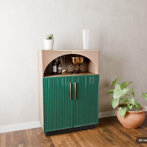 May include: A wooden cabinet featuring a light wood upper section with an arched open shelf and a lower section with two dark green fluted doors. The cabinet has gold vertical handles and contains two wine bottles and two gold goblets.