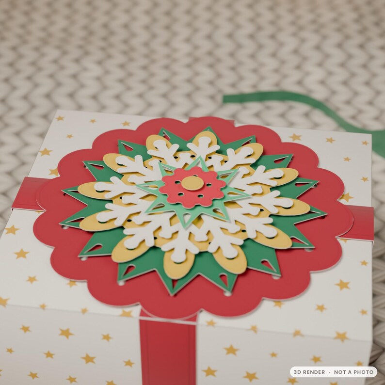May include: A white square gift box decorated with gold stars and a red ribbon. The top features a layered paper ornament with a large red scalloped circle, a green star, a yellow snowflake, a white snowflake, and a small red flower.