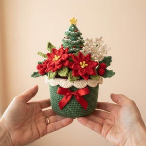 May include: A crocheted Christmas arrangement in a green pot featuring red poinsettia flowers, green leaves, red berries, a white snowflake, and a small green Christmas tree topped with a yellow star, finished with a red satin bow.