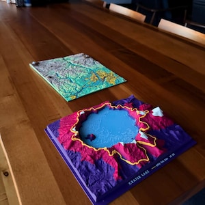 May include: Two square 3D topographic maps. The foreground map depicts Crater Lake in shades of purple, pink, and blue with a yellow rim and the text CRATER LAKE - RIDE THE RIM 33 MI. The background map shows a green and blue landscape.