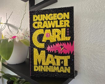 Può includere: Un libro intitolato Dungeon Crawler Carl di Matt Dinniman, con una copertina nera decorata con strass gialli che formano il testo e un grafico rosa di una figura che salta, con strass neri che coprono il resto della superficie.