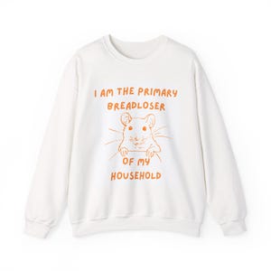 May include: A white crewneck sweatshirt featuring an orange line-art illustration of a mouse peeking over the text I AM THE PRIMARY BREADLOSER OF MY HOUSEHOLD. The graphic is centered on the chest with long sleeves and ribbed cuffs.