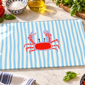 Puede incluir: Una tabla de cortar rectangular de cristal con un patrón de rayas verticales en azul claro y blanco. Centrada en la tabla hay una ilustración estilizada de un cangrejo rojo con puntos naranjas en las pinzas y patas, y dos ojos negros.