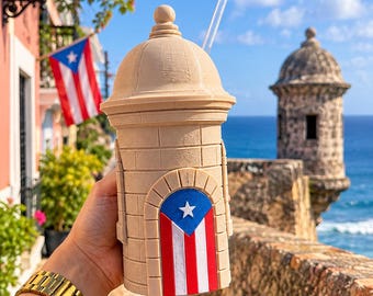 May include: A beige, cylindrical tumbler shaped like a garita sentry box, featuring a textured brick pattern and a prominent arched door decorated with the Puerto Rican flag, complete with a clear straw and a matching domed lid.