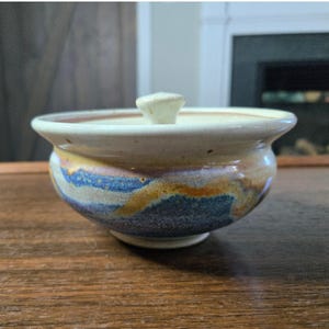 May include: A ceramic bowl with a lid featuring a wide rim and a small knob handle. The exterior displays a mottled glaze pattern with horizontal bands of blue, tan, and cream colors, transitioning into a smooth off-white finish near the top.