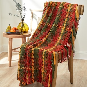 May include: A chunky crochet throw blanket featuring a plaid pattern in shades of deep red, forest green, and mustard yellow. The blanket is finished with long, knotted fringe tassels along the edges and is draped over a wooden chair.