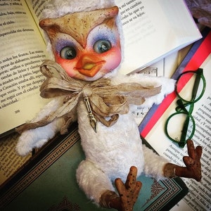Può includere: Un pupazzo gufo bianco con un papillon marrone e beige, occhi blu e un becco marrone. Il pupazzo è seduto su una pila di libri con un paio di occhiali verdi sopra i libri.