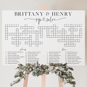 May include: A white sign with a crossword puzzle, featuring the names "Brittany & Henry" and the phrase "sip & solve." The crossword has clues related to the couple. The sign is displayed on a wooden easel, decorated with greenery.