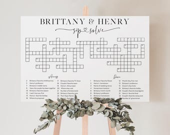May include: A white sign with a crossword puzzle, featuring the names "Brittany & Henry" and the phrase "sip & solve." The crossword has clues related to the couple. The sign is displayed on a wooden easel, decorated with greenery.