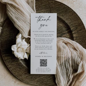 May include: A white rectangular card with the words "Thank you" in elegant script. The card includes a message to family and friends, a QR code, and the names Sierra & Brandon. The card is on a wooden plate with a white flower and beige fabric.