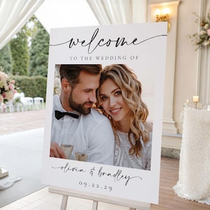 May include: A white wedding welcome sign on an easel. The sign features a photo of a smiling couple and the words "welcome" and "to the wedding of" in elegant script. The names "Olivia & Bradley" and the date "09.22.29" are also displayed.