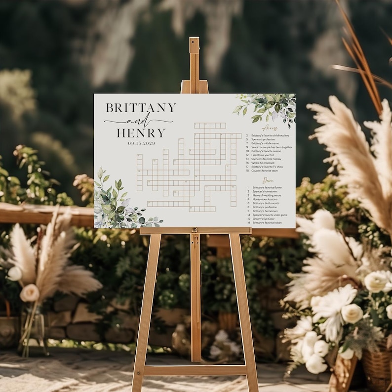 May include: A white wedding sign on a wooden easel featuring a crossword puzzle. The sign includes the names "Brittany and Henry" and the date "09.15.2029", with floral accents. The background is blurred with greenery.