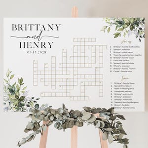 May include: A white wedding crossword puzzle sign with the names "Brittany and Henry" and the date "09.15.2020" in elegant script. The sign is decorated with watercolor greenery and displayed on a wooden easel.