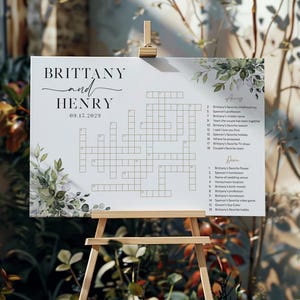 May include: A white wedding crossword puzzle sign on a wooden easel. The sign features the names "BRITTANY and HENRY" with the date "09.15.2029" and a crossword puzzle with clues. Greenery and floral accents frame the top and bottom.
