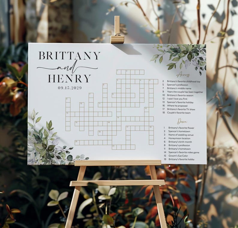 May include: A white wedding crossword puzzle sign on a wooden easel. The sign features the names "BRITTANY and HENRY" with the date "09.15.2029" and a crossword puzzle with clues. Greenery and floral accents frame the top and bottom.