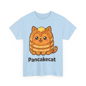 Puede incluir: Una camiseta azul claro con un gráfico de un gato marrón con forma de pila de tortitas, con sirope y un trozo de mantequilla en la cabeza. El gato tiene grandes ojos negros, mejillas rosadas y una boca pequeña. El texto "Pancakecat" está impreso en negro debajo del gato.