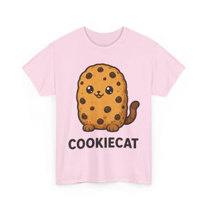 Puede incluir: Una camiseta de manga corta de color rosa claro con un gráfico central de una galleta ovalada de color tostado con chispas de chocolate marrón oscuro, cara de gato, patas y cola. El texto COOKIECAT está impreso en letras negras debajo.