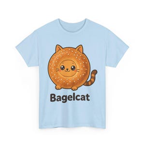 Puede incluir: Una camiseta azul claro con un gráfico de un bagel redondo de color marrón dorado con forma de gato, con semillas de sésamo, orejas pequeñas, una cola a rayas y una cara sonriente. Debajo de la ilustración, el texto Bagelcat está impreso en negro.