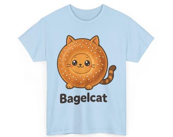 May include: A light blue t-shirt featuring a graphic of a round, golden-brown bagel shaped like a cat with sesame seeds, small ears, a striped tail, and a smiling face. Below the illustration, the text Bagelcat is printed in black.