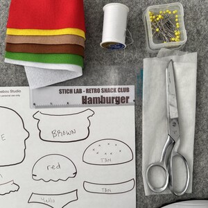 May include: A craft kit for a hamburger featuring stacked red, yellow, brown, green, and white felt sheets, a spool of white thread, a container of yellow-headed pins, metal scissors, and a paper pattern labeled STICH LAB - RETRO SNACK CLUB.
