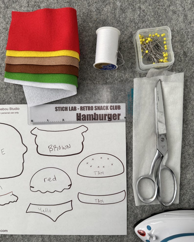 May include: A craft kit for a hamburger featuring stacked red, yellow, brown, green, and white felt sheets, a spool of white thread, a container of yellow-headed pins, metal scissors, and a paper pattern labeled STICH LAB - RETRO SNACK CLUB.