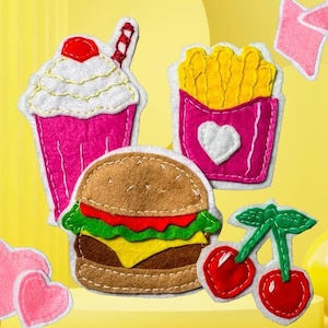 May include: A collection of felt patches featuring a pink milkshake with a red cherry, a carton of yellow french fries with a white heart, a brown hamburger with green lettuce and yellow cheese, two red cherries with green stems, and pink hearts.