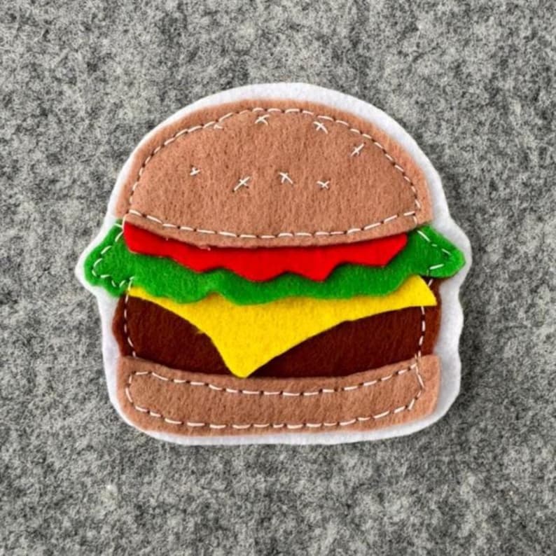 May include: A felt hamburger applique featuring a light brown bun, a red tomato slice, green lettuce, a yellow cheese slice, and a dark brown patty. The layers are held together with white decorative stitching, and the entire piece is outlined in white.