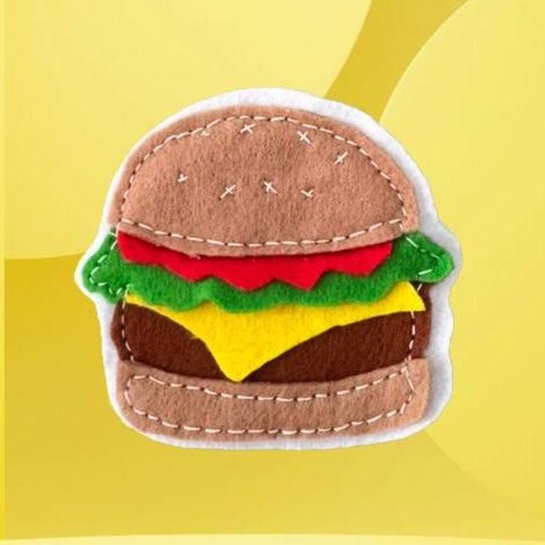 May include: A handcrafted felt hamburger ornament featuring a light brown bun with white stitched details, a dark brown patty, a bright yellow slice of cheese, a wavy green lettuce layer, and a red tomato slice, all assembled with visible white thread.