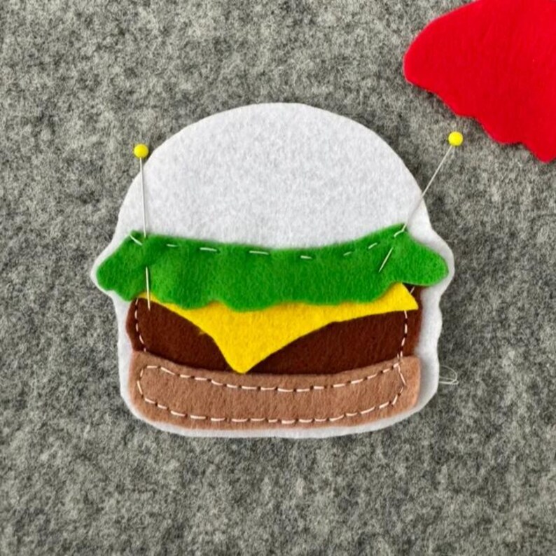 May include: A felt hamburger craft piece featuring a white bun top, a green lettuce layer, a yellow cheese slice, a brown patty, and a tan bun bottom. The layers are held together with white stitching and two yellow-headed sewing pins.