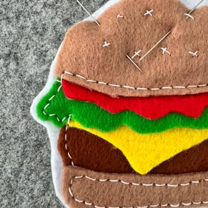 May include: A felt hamburger pincushion featuring layers of brown bun, red tomato, green lettuce, yellow cheese, and a brown patty. The layers are joined with white decorative stitching. A yellow-headed pin and silver needles are inserted.