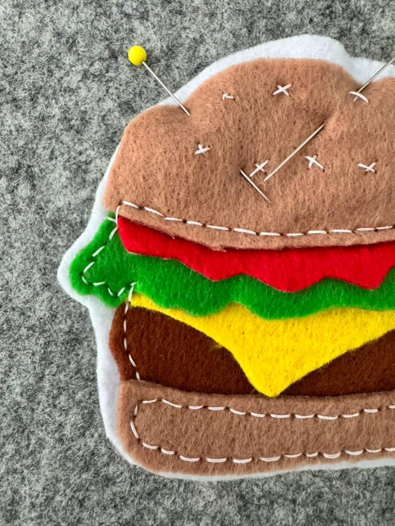 May include: A felt hamburger pincushion featuring layers of brown bun, red tomato, green lettuce, yellow cheese, and a brown patty. The layers are joined with white decorative stitching. A yellow-headed pin and silver needles are inserted.