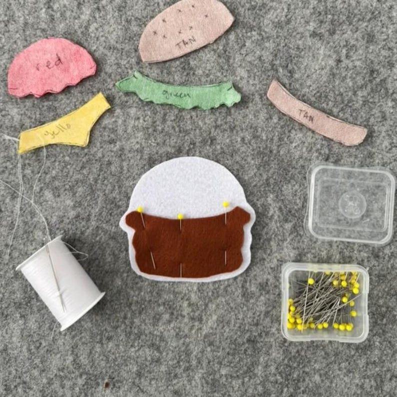 May include: A craft project featuring a white and brown felt cupcake shape pinned with yellow needles, surrounded by loose felt pieces in pink, tan, green, and yellow labeled with text, a spool of white thread, and a clear plastic box of yellow pins.