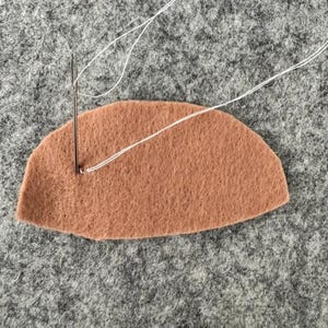 May include: A semi-circular piece of tan felt fabric being stitched with a silver sewing needle and a thin white thread. The needle is positioned vertically, piercing the felt near the left edge, with the thread extending outward.