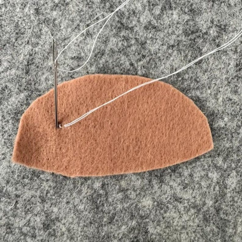 May include: A semi-circular piece of tan felt fabric being stitched with a silver sewing needle and a thin white thread. The needle is positioned vertically, piercing the felt near the left edge, with the thread extending outward.