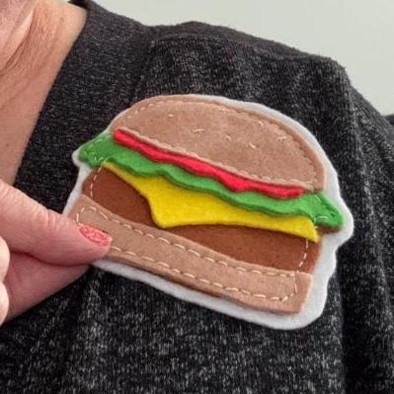 May include: A felt hamburger patch featuring a tan bun, a brown patty, a yellow slice of cheese, a red layer, and a green lettuce layer. The layers are stitched together with white thread and mounted on a white felt backing on a dark gray fabric.