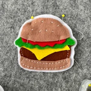 May include: A felt hamburger ornament featuring a light brown bun, a dark brown patty, a bright yellow cheese slice, a red tomato layer, and green lettuce. The layers are held together with white stitching and two yellow-headed sewing pins.
