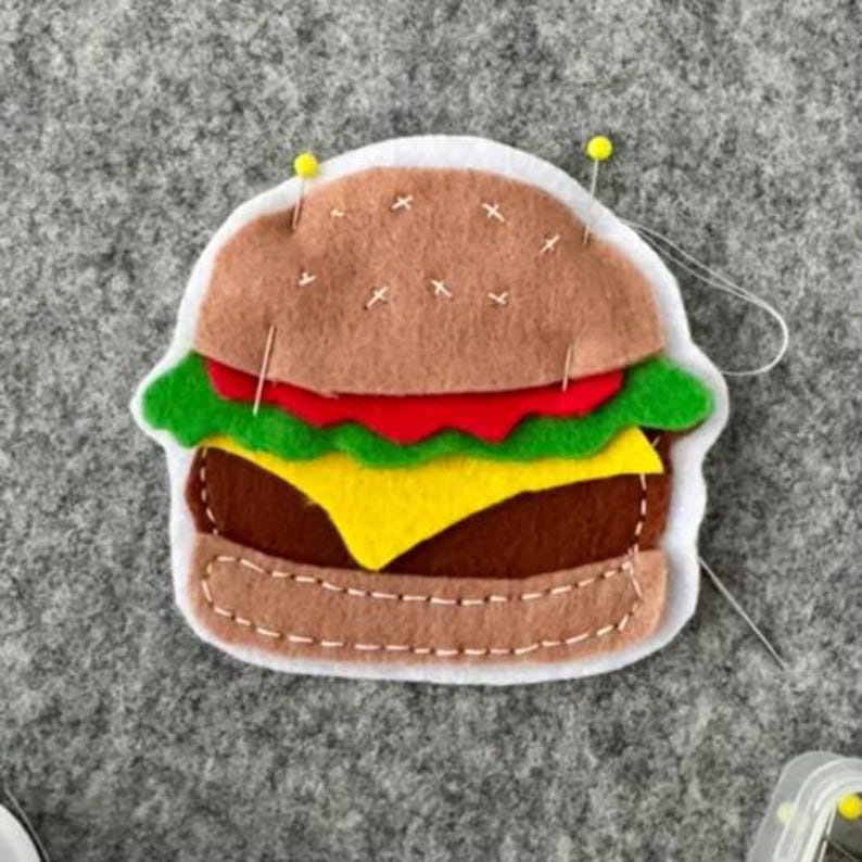 May include: A felt hamburger ornament featuring a light brown bun, a dark brown patty, a bright yellow cheese slice, a red tomato layer, and green lettuce. The layers are held together with white stitching and two yellow-headed sewing pins.