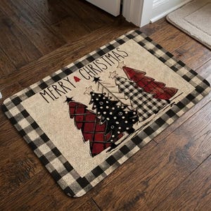 May include: A rectangular doormat with a beige textured center and a black and white buffalo check border. The mat features the text MERRY CHRISTMAS and a graphic of four stylized Christmas trees in red, black, and white patterns.