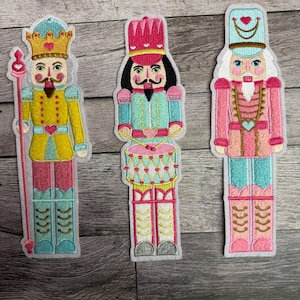 Könnte beinhalten: Drei gestickte Nussknacker-Aufnäher in leuchtenden Farben. Die linke Figur trägt eine goldene Krone und hält einen Stab. Die mittlere Figur spielt eine Trommel mit rosa und blauen Akzenten. Die rechte Figur trägt einen hellblauen Hut und Uniform.