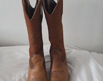 May include: A pair of tan leather cowboy boots featuring a mid-calf height, pointed toes, and decorative stitching on the vamps and shafts. The boots have pull tabs at the top openings and a stacked heel design, showcasing a classic western silhouette.