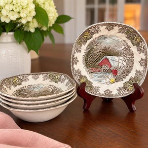 May include: A set of ceramic bowls and a plate featuring a winter scene with a red covered bridge. The rim of each piece is decorated with a brown and green leaf pattern. One plate is displayed upright on a dark wooden stand.
