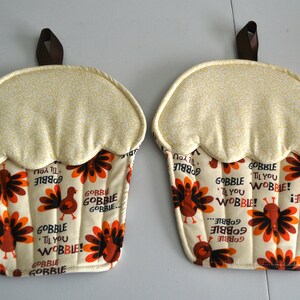 May include: Two cupcake-shaped pot holders. The top is a light yellow fabric with a small pattern. The bottom has a cream background with turkeys and the words "Gobble 'Til You Wobble!" in orange and brown. Each has a brown loop.
