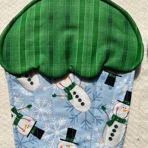 May include: A cupcake-shaped pot holder featuring a green striped top section and a light blue bottom section decorated with a repeating pattern of snowmen wearing black top hats and green scarves, accented by white snowflakes and a hanging loop.