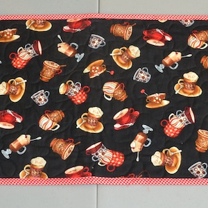 May include: A black quilted table runner with a red and white checkered border. The runner features a repeating pattern of coffee cups and mugs.