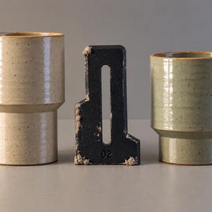 May include: Two cylindrical ceramic cups with a stepped base design, one in a speckled cream finish and the other in a speckled sage green finish, positioned on either side of a black metal object with the number 02 embossed on it.
