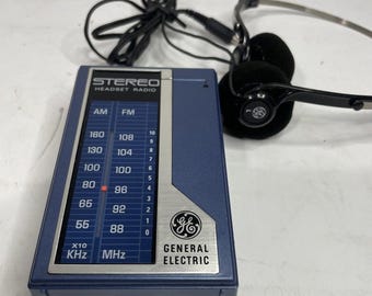 Può includere: Una radio con cuffie stereo General Electric blu con un pannello di controllo argentato che mostra le scale di frequenza AM e FM. Il dispositivo include cuffie nere cablate con cuscinetti in schiuma e il logo GE sul case.